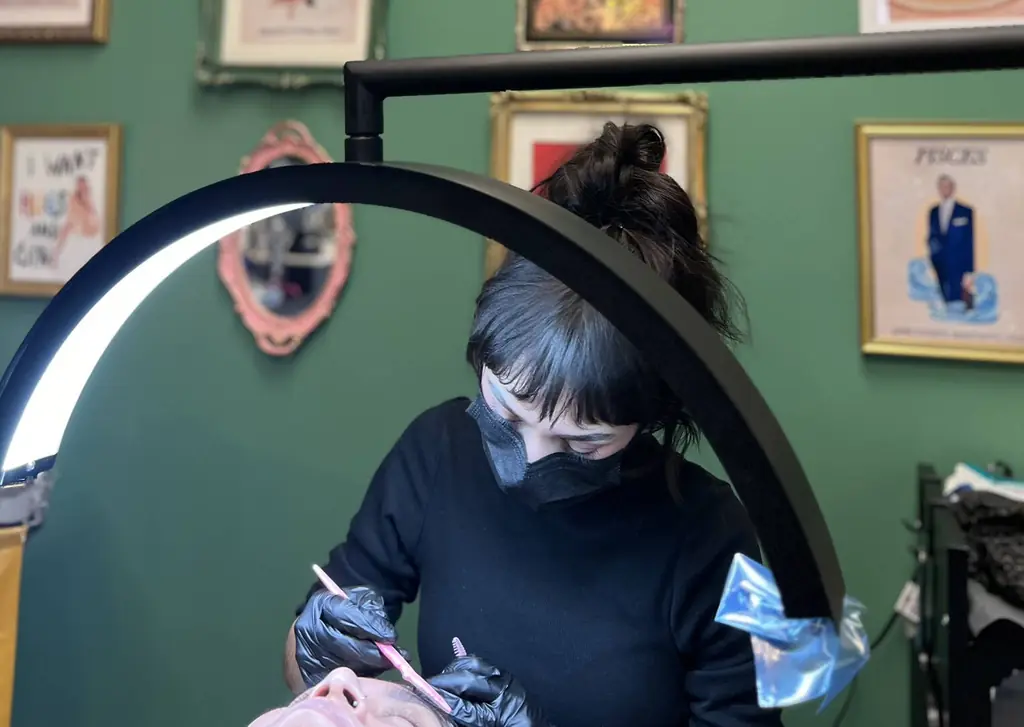 Katie, in a mask and gloves, working on a client's brows under a ring light in the green-walled suite